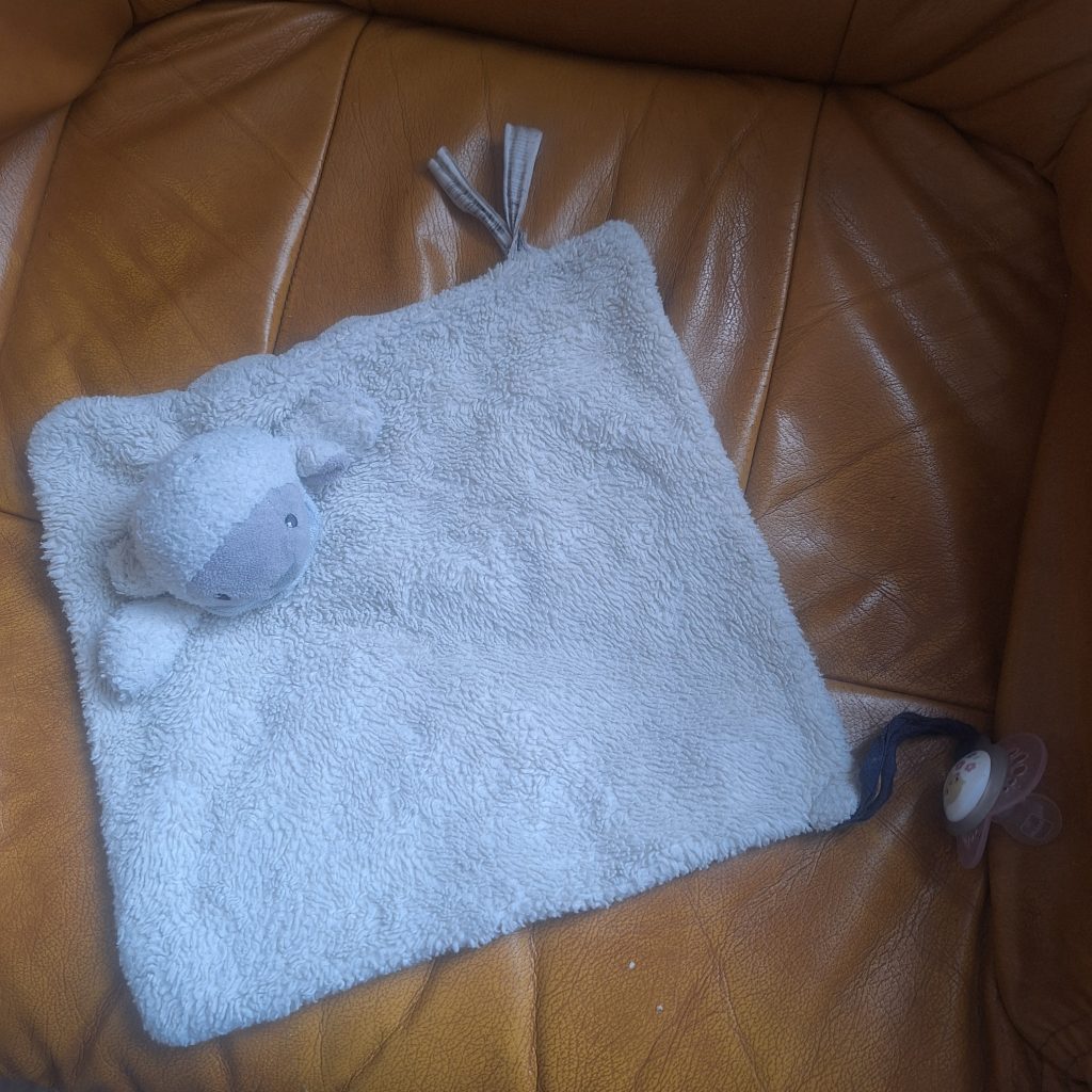Doudou mouton blanc avec étiquettes doubles, posé sur un fauteuil en cuir marron.